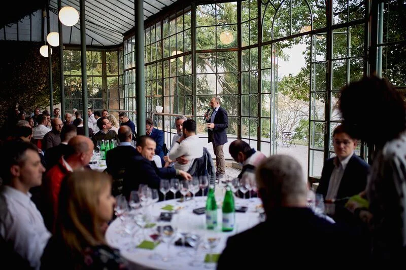 Persone sedute a dei tavoli che degustano vino in una bellissima location con grandi vetrate