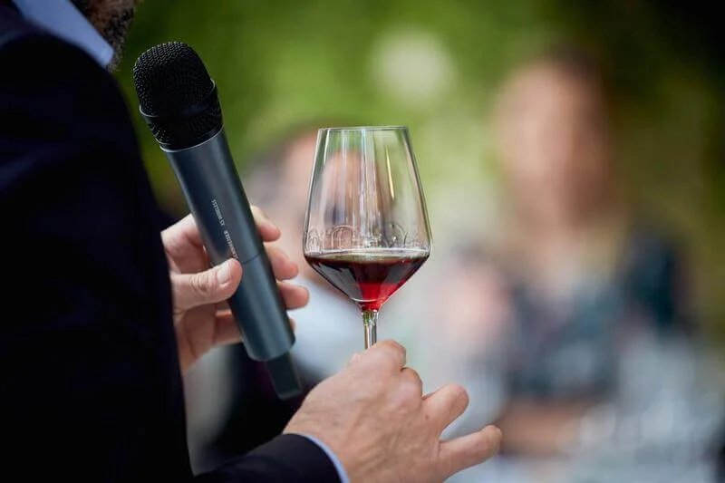 Persona in giacca e camicia, che tiene in mano un bicchiere di vino e un microfono