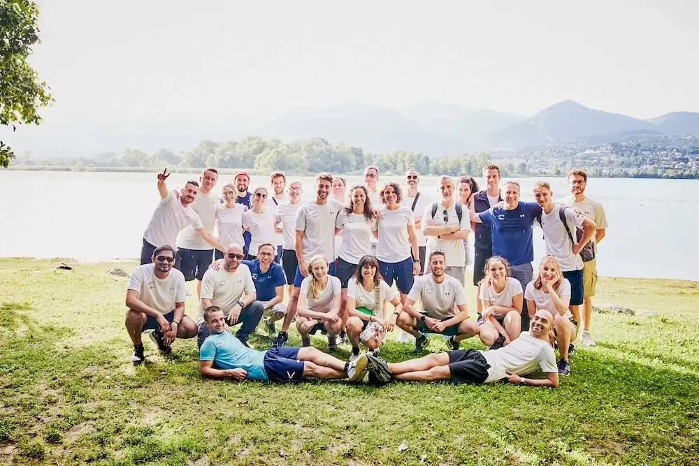 Team aziendale sorridente durante un'attività di team building sul lago in Lombardia, con sfondo montano e atmosfera collaborativa.