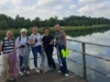 Gruppo aziendale sorridente posa con i sacchi raccolti dopo una giornata di team building ecologico e solidale al Parco Nord, con vista sul lago.