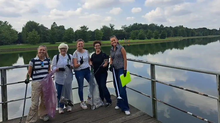 Gruppo aziendale sorridente posa con i sacchi raccolti dopo una giornata di team building ecologico e solidale al Parco Nord, con vista sul lago.