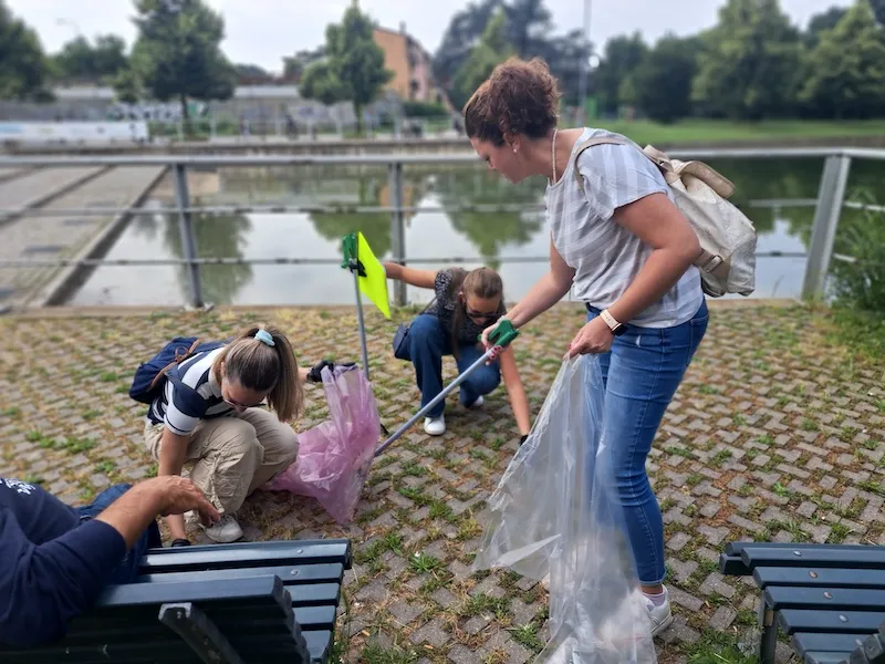 Attività di team building solidale con volontari che raccolgono rifiuti vicino a una panchina in un’area verde.